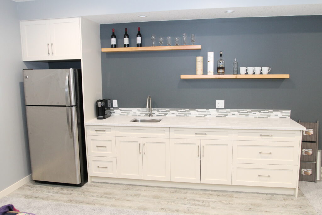 Finished basement wet bar with white cabinets, floating wood shelves and a stainless fridge
